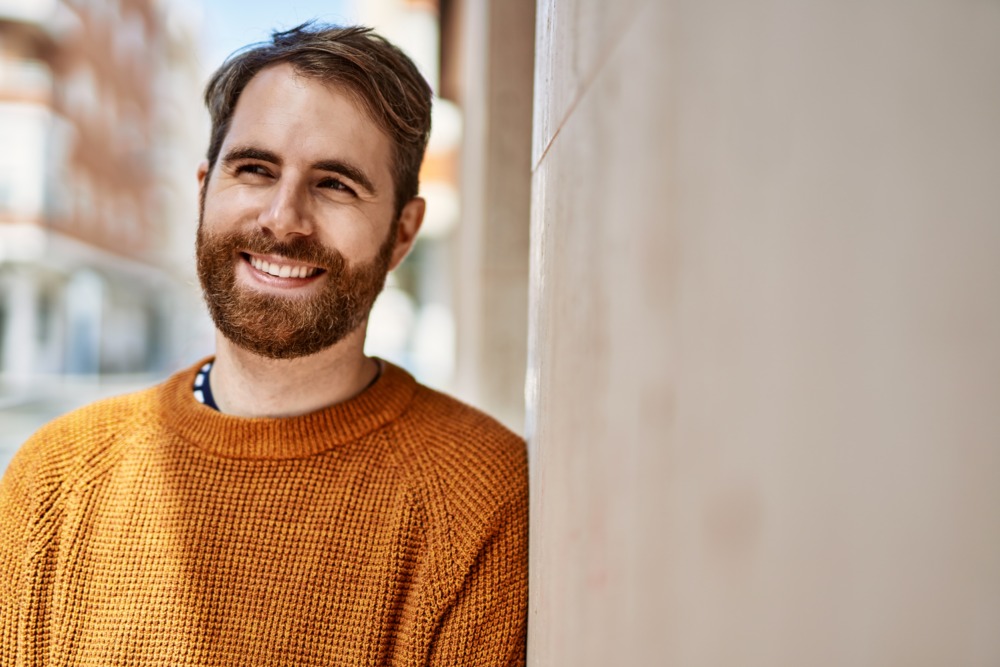 Young man with a beard smiling and leaning against a wall outside in the sunshine.
