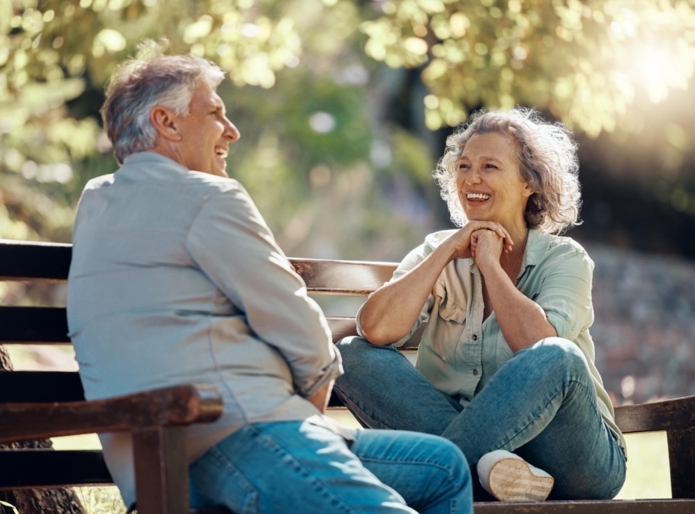 A man and woman sit on a park bench, facing each other and laughing in conversation.