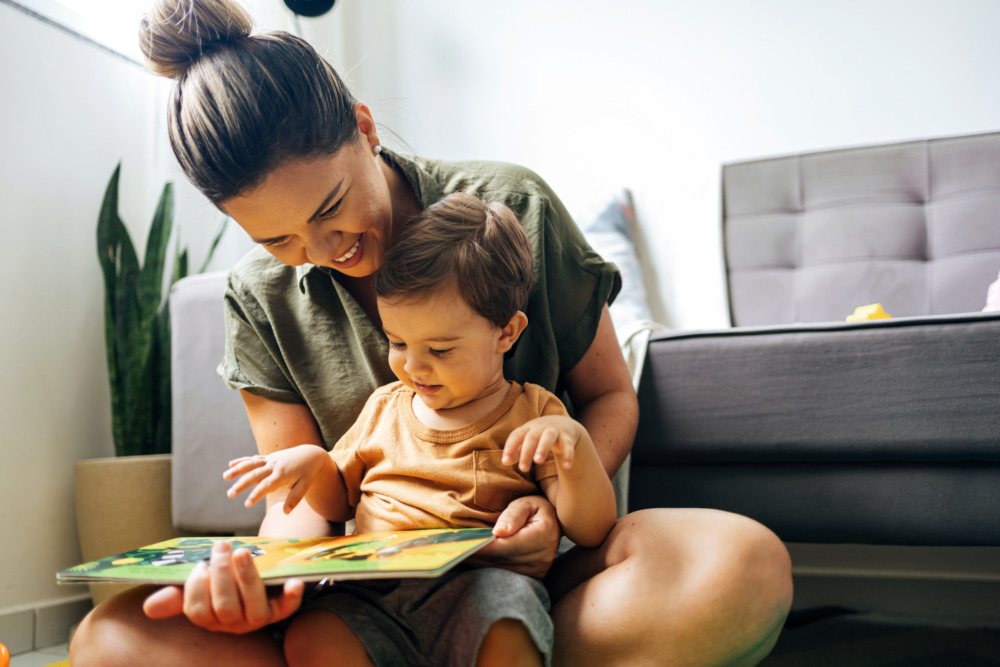 A mother sat on the floor with her young son on her lap, reading a book together and laughing.