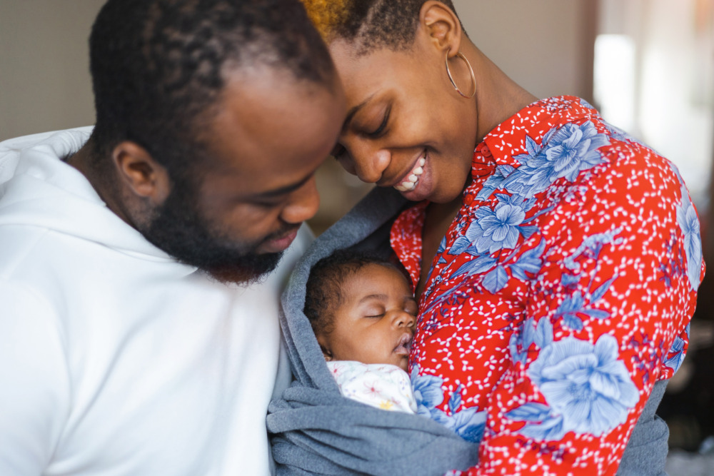 A mother and father smiling at their new born baby, swaddled between them.