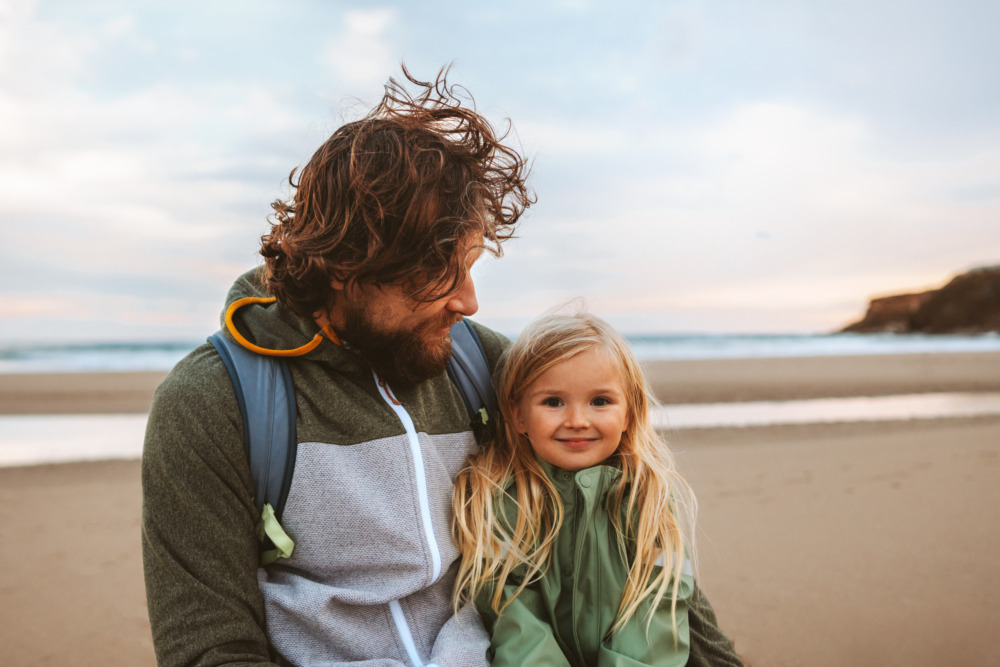 Father with his young daughter on the beach. He looks at her, she looks at the camera and smiles.