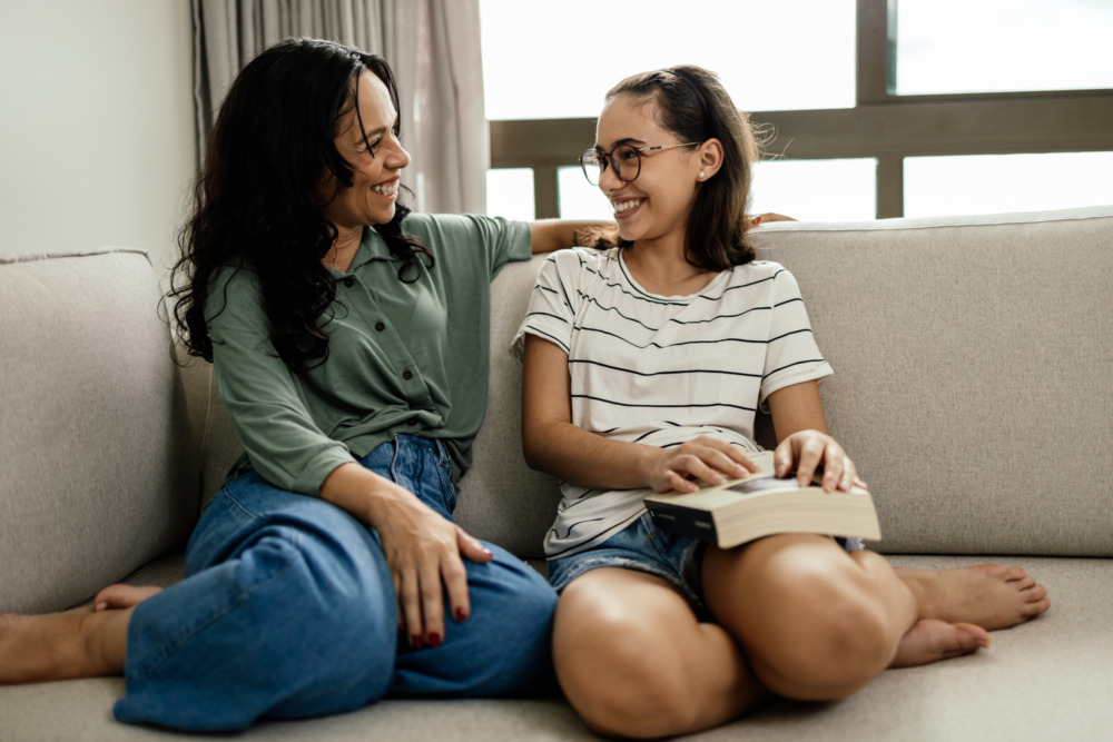 A mother and her teenage daughter laughing happily on the sofa, the girl holding a book on her leg.
