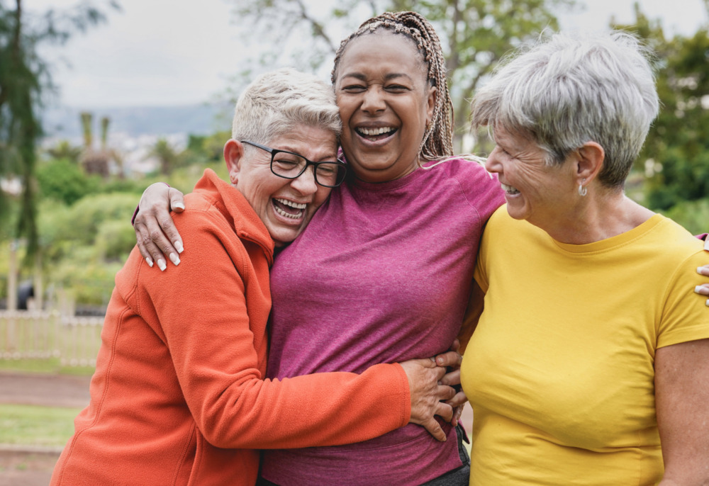 Group of three elderly women laughing and hugging in a park.