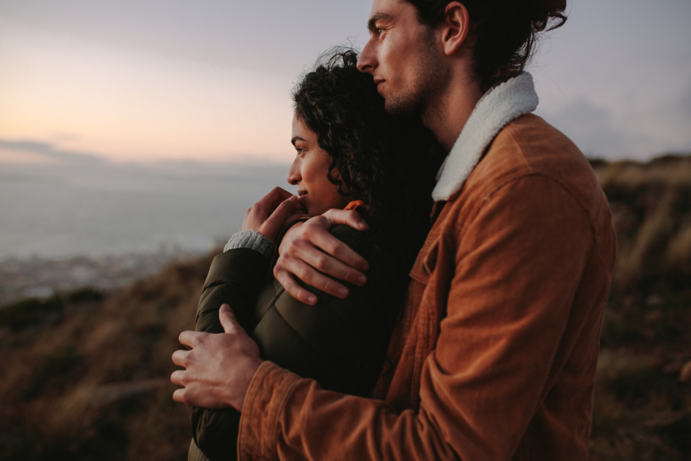 Young couple romantically looking out over the ocean at sunset. The man hugs the woman from behind.