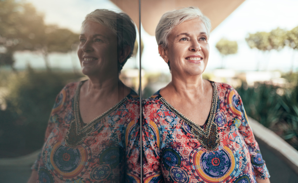 A grey haired, middle aged woman looking out into the distance with her reflection in a window to the left.