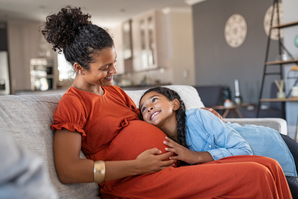 A woman sits on the sofa with her hand rested on her pregnant belly. A young child cuddles into her, smiling up at her.