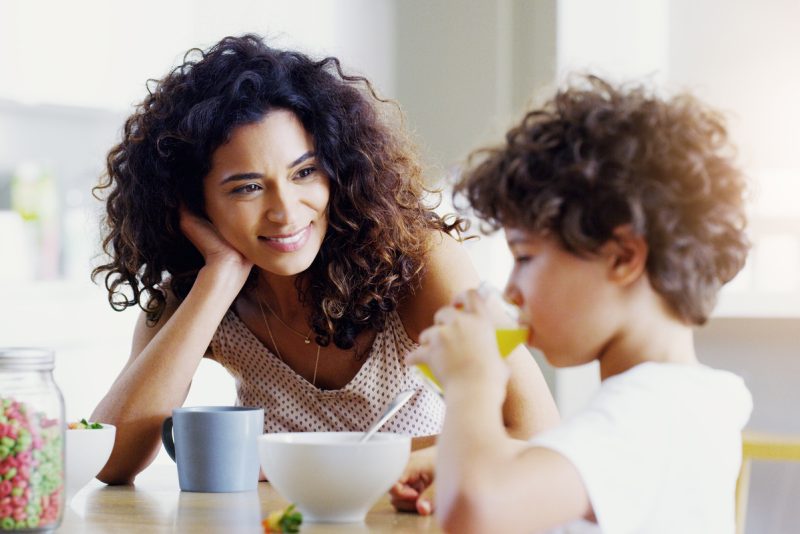 Woman with curly brown hair smiling at her young son drinking orange juice
