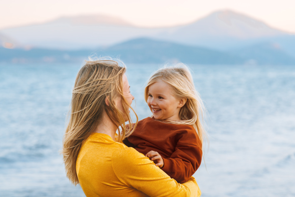 A mother with her back to the camera holding her daughter in her arms. They face each other, with the ocean in the background.