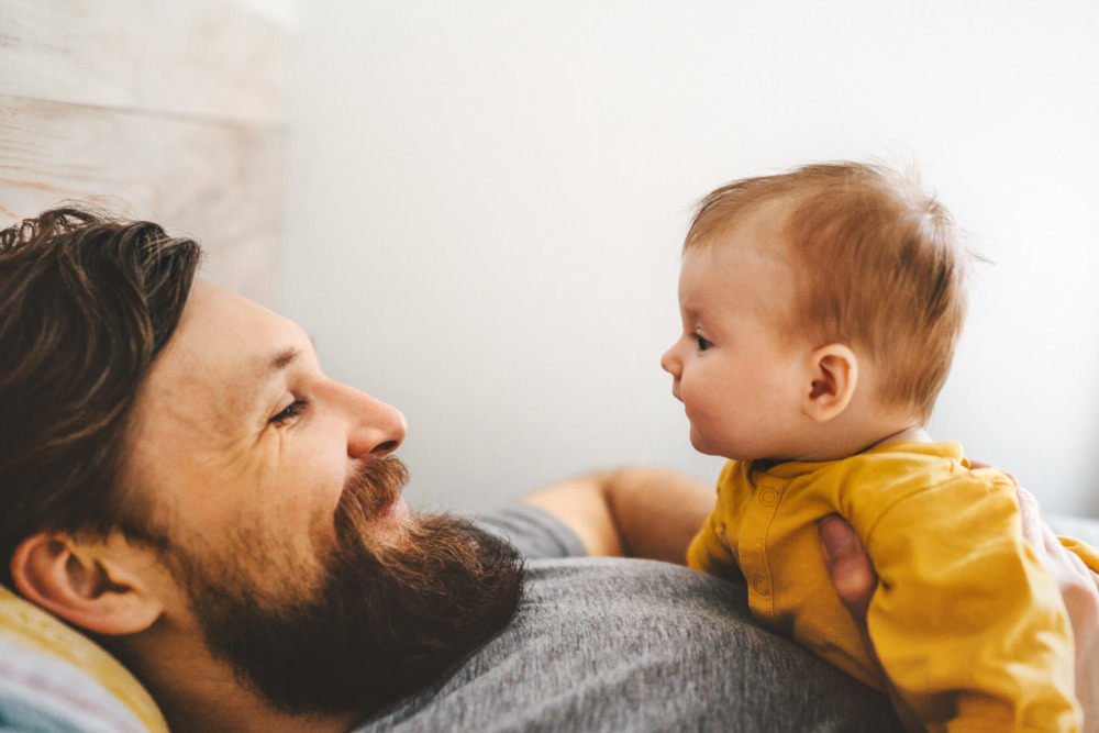 A father lying on his back, smiling at his young baby, who's lying on his chest and looking at him.
