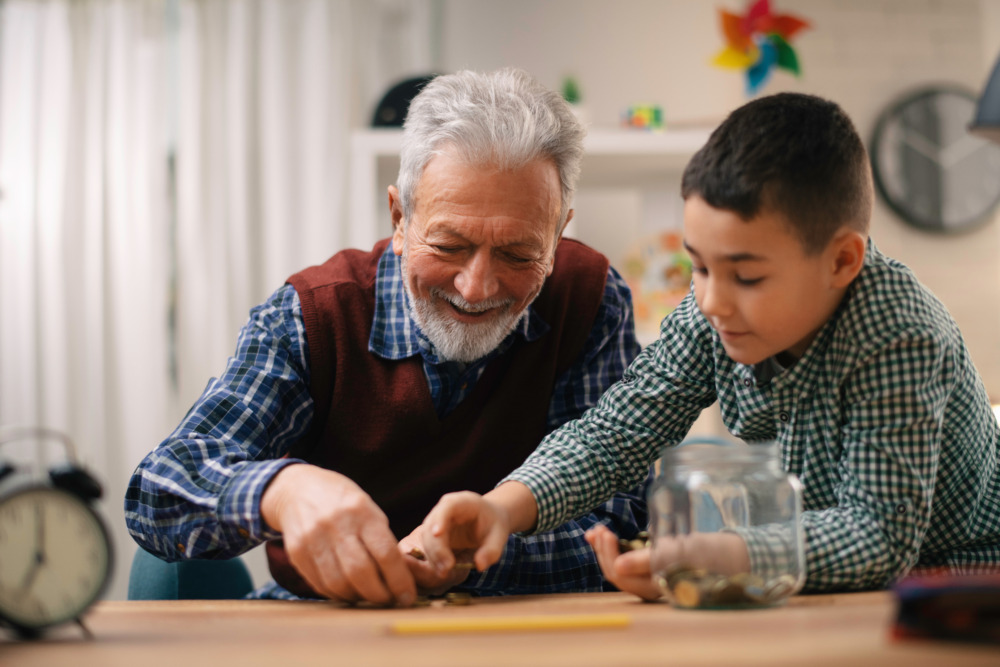 A grandfather and his grandson sit at a table, counting coins into a glass bottle, and smiling together.