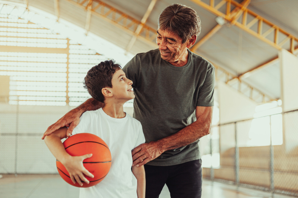Grandfather and grandson smiling at each other on a basketball court. The grandson holds a basketball in one arm.