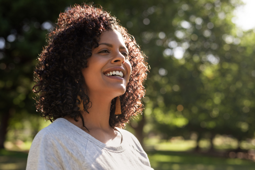 A close up of a woman, standing outside and smiling in the sunshine in a park.
