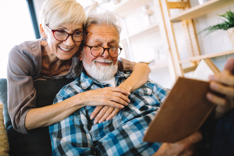 An older woman stands behind her husband, who is reading a book, embracing him with her arms around his shoulders.