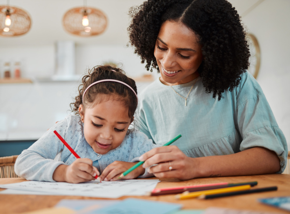 A young mother and her daughter, colouring a picture together at a table inside.