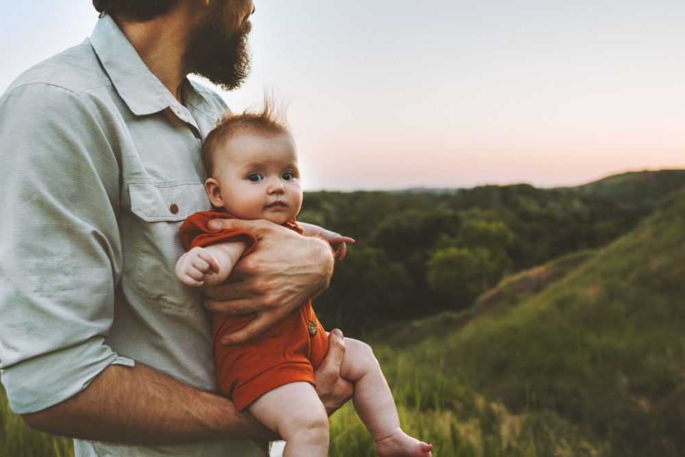 Close up of a baby, being held by its father who is walking outdoors and looking away.