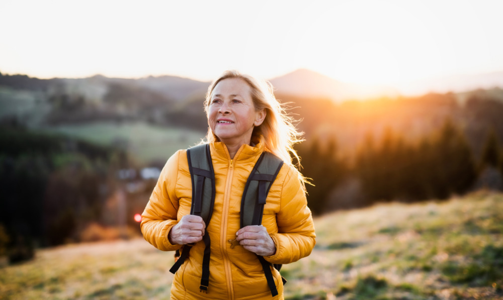 Woman wearing a puffer coat and backpack, hiking through the mountains as the sun comes up behind her.