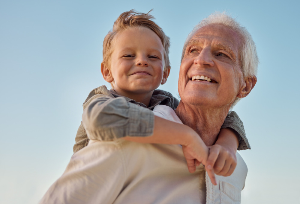 A grandfather is giving his young grandson a piggyback. They both smile, with a bright blue sky behind them.
