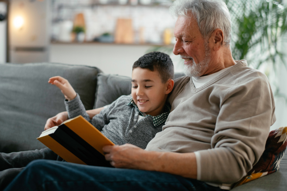 A grandfather and his grandson sit on a sofa, smiling and reading a book together.