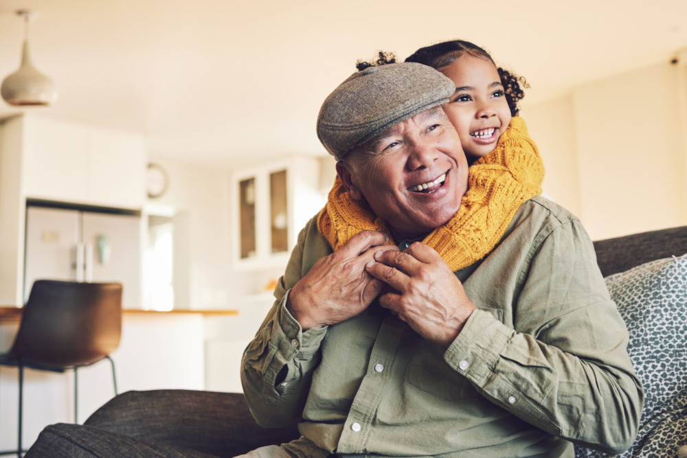 A grandfather sat on a sofa laughing, while his young granddaughter hugs him around the neck from behind.