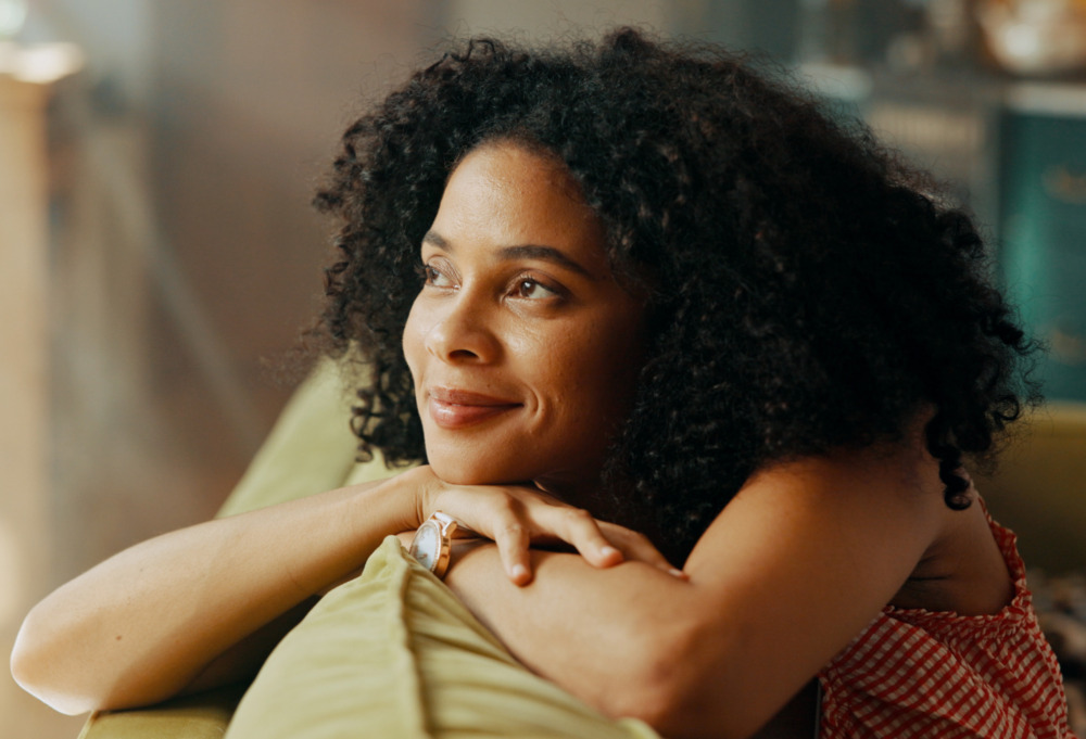Smiling woman with dark, curly hair on couch at house.
